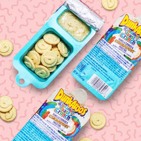 An opened snack tub of vanilla flavored Dunkaroo biscuits with cinnamon toast crunch frosting flavor dip on a light pink patterned background.